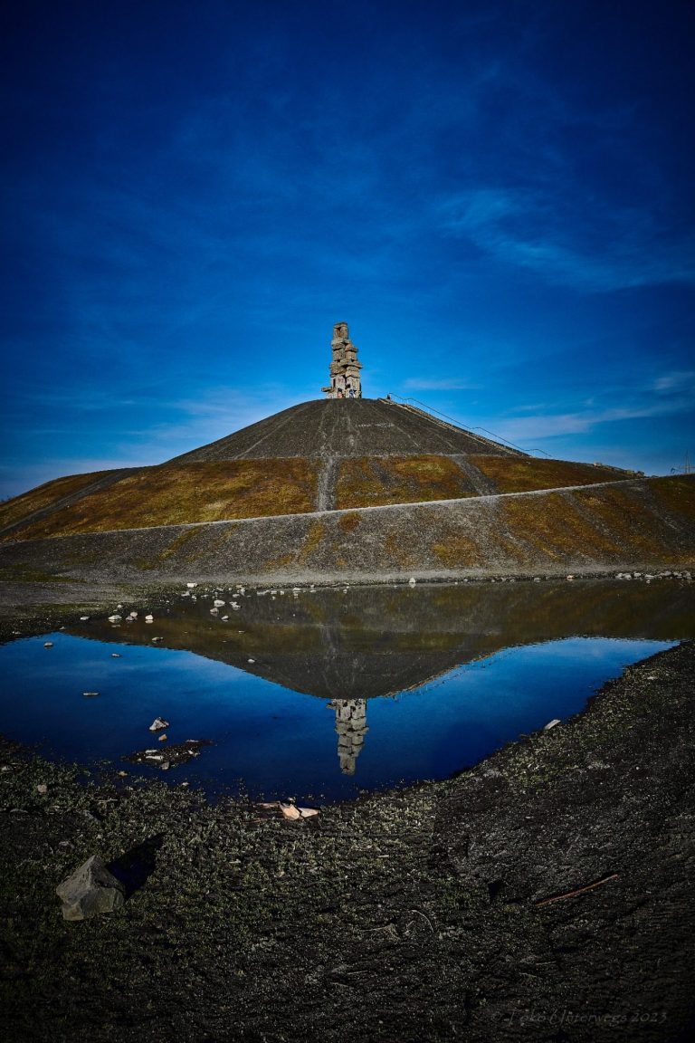 Steinerne Statue auf einem Hügel, reflektiert im ruhigen Wasser eines Teiches.