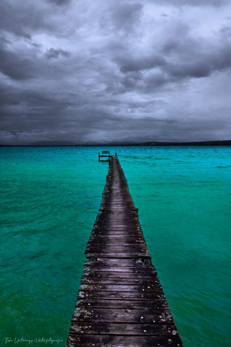 Ein Holzsteg führt über das turquoise Wasser unter einem bewölkten Himmel.