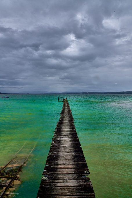 Holzsteg führt über türkisfarbenes Wasser unter bewölktem Himmel.