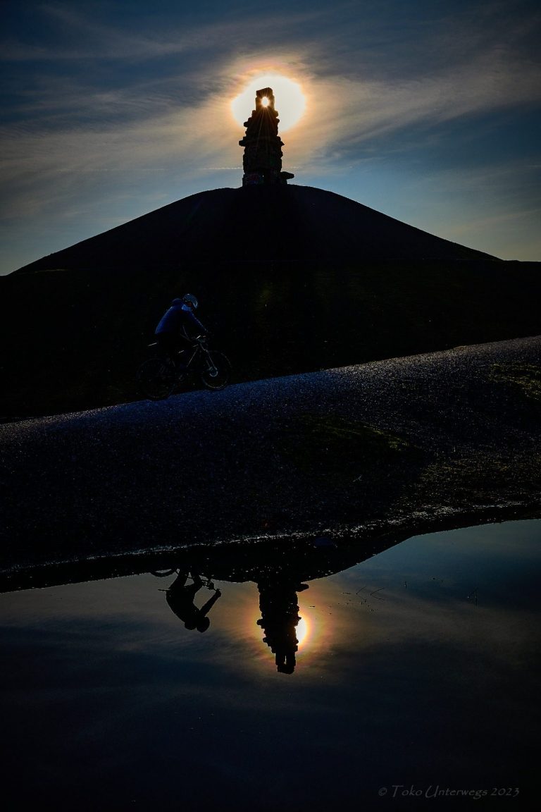 Sonnenuntergang hinter einem Hügel mit einem silhouettierten Turm und Spiegelung im Wasser.